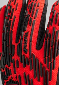 Czerwone rękawice z fakturowanym czarnym gumowym gripem na dłoni. Materiał to tkanina z siateczkowym pokryciem, z wzmocnionym obszarem nadgarstka i detalem logo.