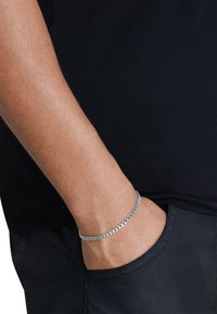 Sølvkjede armbånd på håndleddet, med en polert overflate og sammenflettede lenker. Båret over en svart skjorte nær en mørk lomme.