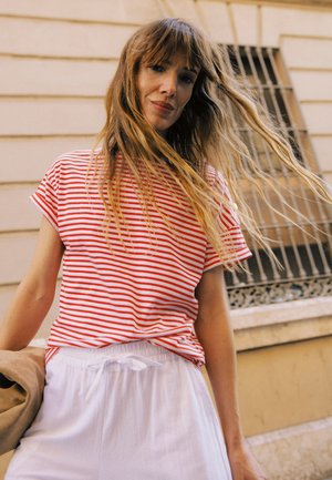 Jeune femme aux longs cheveux portant une chemise rouge et blanche à rayures et un pantalon blanc, se tenant à l'extérieur près d'un bâtiment beige avec des fenêtres grillagées.
