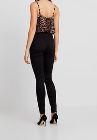 Kvinna i leopardmönstrad ärmlös topp, svarta högmidjade skinny jeans och svarta högklackade skor, stående mot en neutral bakgrund.
