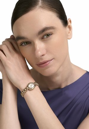 Vrouw met groene ogen en donker haar draagt een goudkleurige armbandhorloge met kralen, legt haar handen dicht bij haar gezicht en draagt een marineblauw topje.