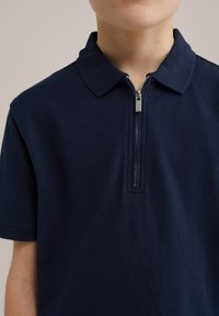 Polo de culoare bleumarin, cu un material texturat, având un guler și un fermoar argintiu în față pentru închidere.