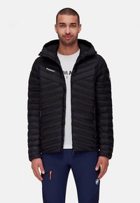 Άνδρας φοράει μαύρο καπιτονέ μπουφάν με κουκούλα, λευκό μπλουζάκι με εκτύπωση και ναυτικές μπλε παντελόνες με φερμουάρ στις τσέπες, στέκεται στραμμένος μπροστά.