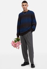 Gebreide trui met blauwe en bruine strepen, grijze broek en zwarte schoenen. Houdt roze lelies vast met zwarte accenten op de bloemen.