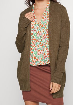 Femme portant un long cardigan marron sur une chemise à boutons à motifs floraux et une jupe couleur rouille, une main ajustant le col du cardigan.