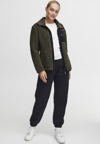 Olijfgroene fleecejack met een zwarte rits aan de voorkant en kraag. Gekleed met zwarte sweatpants en witte sneakers, wat een casual uitstraling geeft.