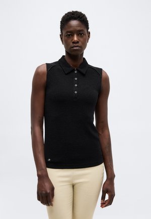 Γυναίκα που φοράει μαύρο αμάνικο πουκάμισο με γιακά και μπεζ παντελόνια, στέκεται μπροστά σε απλό ανοιχτό φόντο, κοιτάζοντας μπροστά.