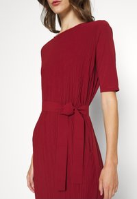 Rotes, knielanges Kleid aus strukturiertem Stoff, mit kurzen Ärmeln und einem gebundenen Taillengürtel. Mit einem Boots-Ausschnitt und sanften Falten versehen.