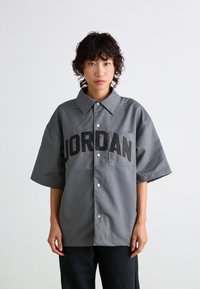 Gray short-sleeve shirt with a pointed collar, featuring a large black "JORDAN" logo on the front and a chest pocket. Smooth fabric texture.
