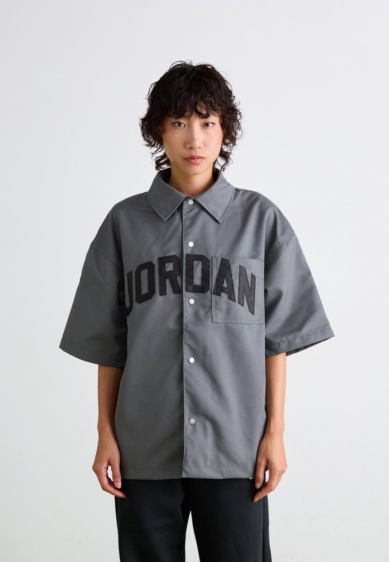 Gray short-sleeve shirt with a pointed collar, featuring a large black "JORDAN" logo on the front and a chest pocket. Smooth fabric texture.