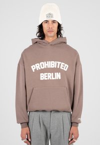Persona che indossa un hoodie marrone con il testo "PROHIBITED BERLIN", pantaloni grigi a pieghe, un beanie bianco e anelli d'argento sulle dita.