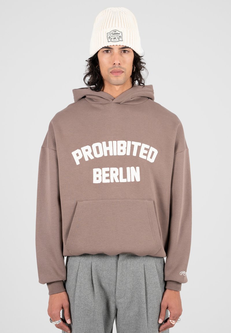 Persona che indossa un hoodie marrone con il testo "PROHIBITED BERLIN", pantaloni grigi a pieghe, un beanie bianco e anelli d'argento sulle dita.