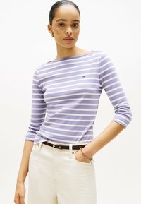Lila-weiß gestreiftes Langarmoberteil mit Bootsausschnitt und schmaler Passform, kombiniert mit hellen Hosen und einem schwarzen Gürtel.