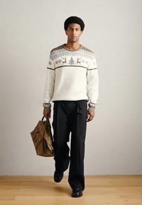 Λευκό πουλόβερ με σχέδιο τάρανδων και νιφάδων χιονιού, σε συνδυασμό με μαύρο παντελόνι. Κρατάει ένα καφέ τσάντα με υφασμάτινη υφή.