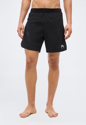 Férfi modell fekete sportos rövidnadrágot visel, perforált oldalpanelek és a bal lábszáron egy kis logó látható, mezítláb áll egy egyszerű háttér előtt.