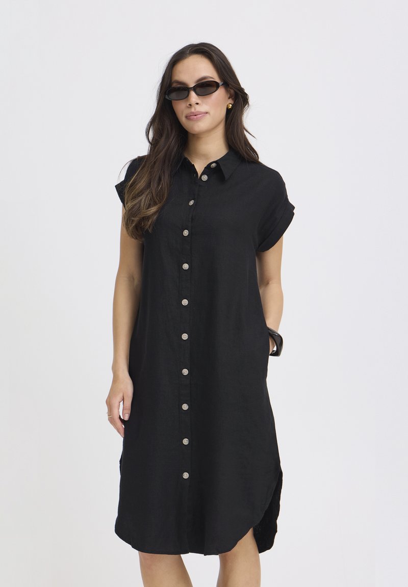 Femme portant une robe noire à manches courtes avec boutons jusqu'aux genoux et des lunettes de soleil noires, debout devant un fond clair uni.