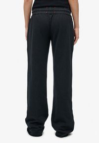 Persoon in een losse zwarte jogginsbroek en een zwart topje, gezien vanuit de achterkant tegen een effen witte achtergrond.