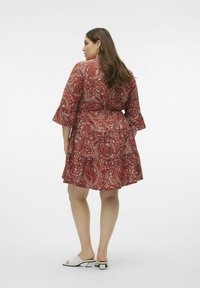 Robe paisley rouge avec de longues manches en forme de cloche, ceinture à la taille et jupe en couches. Associée à des talons blancs à bout ouvert sur un fond neutre.