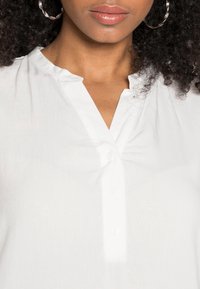 Blusa blanca con escote en V y pequeños pliegues en el cuello, con una tapeta de botones y tela suave y ligera.