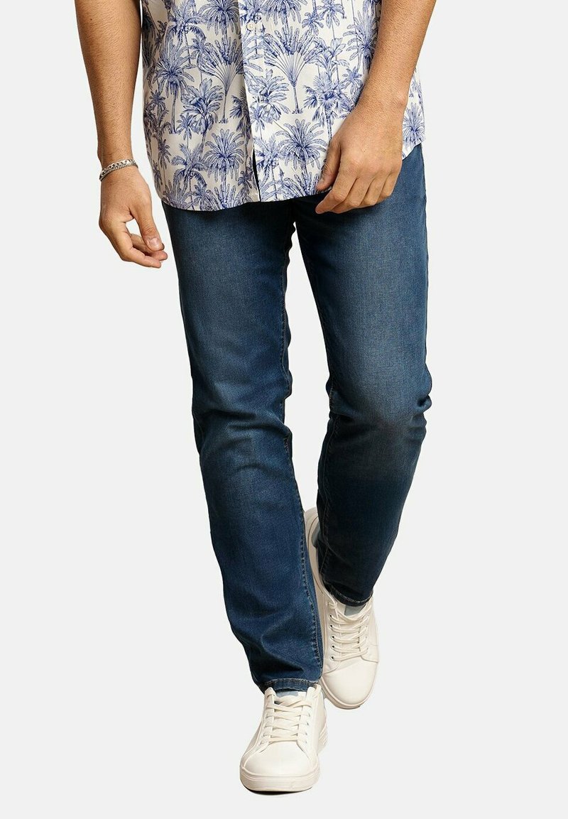 Conjunto casual que incluye una camisa azul con estampado, jeans azul oscuro y zapatillas blancas. La camisa tiene un diseño de hojas de palma y mangas cortas.