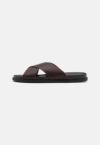 Sandale à glissière à lanières croisées de couleur marron, dotée d'une finition mate, d'une semelle noire plate et d'une texture lisse. Design minimaliste sans matériel visible.