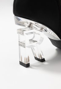 Chaussure noire avec un talon transparent en forme de lettre "R" et embouts en caoutchouc noir sur une surface blanche.