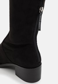 Botina neagră din suede, cu înălțime până la genunchi, cu fermoar lateral, textură netedă și toc masiv negru. Prezintă un zipper pull cu marcă.