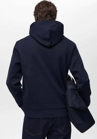 Marinblå huvtröja med texturerad yta, försedd med en känguruficka och dragsko i huvan. Kombineras med mörka denimjeans.