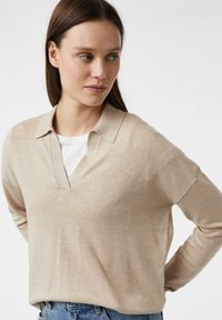 Beżowy sweter z dzianiny z dekoltem w kształcie litery V i miękką teksturą. Posiada kołnierz i luźny krój, zestawiony z niebieskimi dżinsami dla kontrastu.
