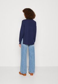 Person med lockigt hår som bär en marinblå tröja, ljusblå vida jeans och bruna klackklossar, stående mot en vit bakgrund.