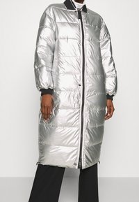 Sølvopustet frakke med en blank, quiltet overflade, sorte accenter, en frontlynlås og elastiske manchetter; knælangt design med en afslappet pasform.