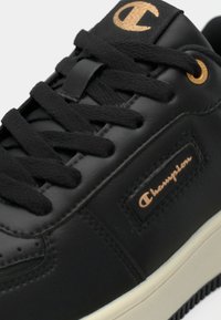 Μαύρο δερμάτινο αθλητικό παπούτσι με μαύρα κορδόνια, χρυσό λογότυπο Champion στη γλώσσα και πλευρά, και λευκή ελαστική σόλα σε κοντινή λήψη.