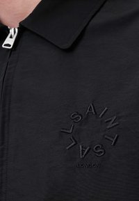Čierna bunda s predným zipsom, s plochým golierom. Vyšívané kruhové logo zodpovedajúcou čiernou niťou, s nápisom "SAINT LONDON."