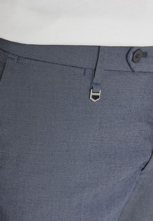 Primer plano de la pretina de unos pantalones azules con textura, con un botón negro, trabillas y un detalle decorativo de cierre plateado.