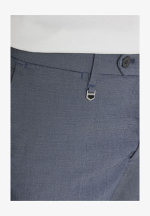 Primer plano de la pretina de unos pantalones azules con textura, con un botón negro, trabillas y un detalle decorativo de cierre plateado.