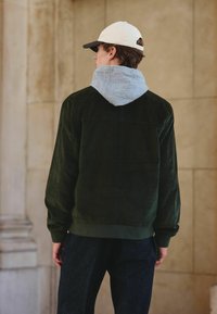 Groene corduroyjas met grijze capuchon, met ribbels aan de mouwen en onderkant, gedragen over donkere broeken, van achter bekeken in een neutrale omgeving.