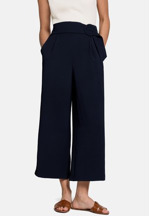 Femme portant un pantalon noir court à taille haute, jambes larges, avec un détail de ceinture, un haut blanc sans manches et des sandales marron à enfiler.