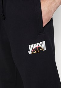 Detailní záběr černých kalhot s rukou v kapse a bílým logem "JORDAN" nad stylizovaným basketbalovým motivem.