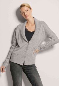 Lichtgrijze cardigan van een zachte stof, met een sjalkraag, knopen aan de voorkant en twee voorzakken. Gecombineerd met een zwarte top en donkere jeans.