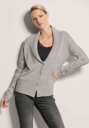 Hellgrauer Cardigan aus weichem Material, mit einem Schalkragen, vorderen Knöpfen und zwei vorderen Taschen. Kombiniert mit einem schwarzen Oberteil und dunklen Jeans.