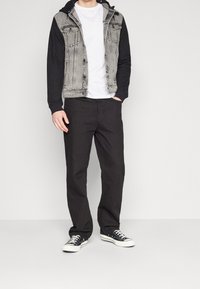 Man som bär en svart och grå denimjacka med svarta ärmar, vit t-shirt, svarta byxor och svarta låga sneakers som står mot en vit bakgrund.