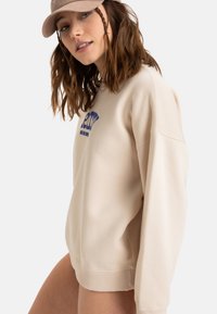 Beige Sweatshirt mit dropped Schultern, mit einem blauen Grafiklogo auf der Vorderseite. Das Material scheint weich zu sein und hat gerippte Bündchen und Saum.
