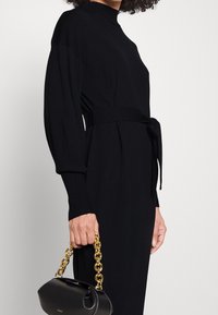 Robe tricotée noire à manches longues, col haut et taille nouée. Présente des poignets côtelés. Sac à main noir brillant avec une anse en chaîne dorée.