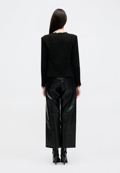 Žena stojaca chrbtom k divákovi, oblečená v čiernom textúrovanom tričku s dlhým rukávom, širokých čiernych kožených nohaviciach a čiernych vysokých čižmách na podpätku.