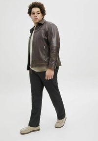 Brown leather jacket with a zippered pocket, grey shirt underneath, black pants, and beige slip-on shoes. Model shown in a standing pose.