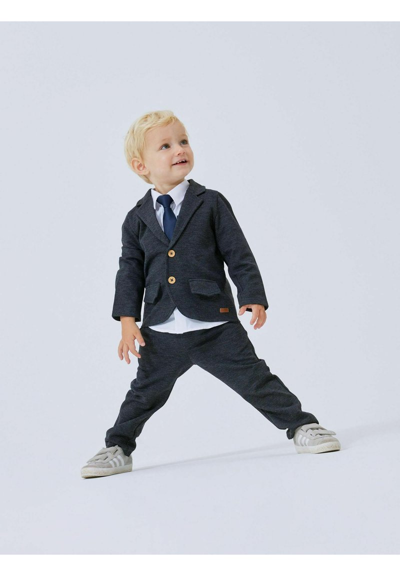 Completo grigio scuro con bottoni, pantaloni abbinati, camicia bianca e cravatta blu. Tessuto morbido, design su misura e scarpe da ginnastica leggere completano il look.