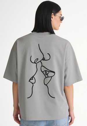 Personne aux cheveux noirs porte un t-shirt gris oversize avec un art linéaire abstrait noir de visages dans le dos, associé à un jean bleu clair.