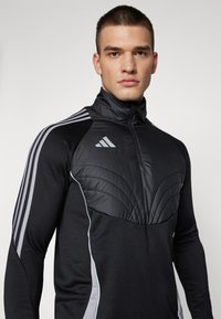 Pull-over de sport noir avec col haut, design à l'avant matelassé et accents argentés. Comprend trois bandes verticales sur les épaules.