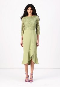 Robe mi-longue de couleur vert clair, présentant un corsage en dentelle avec des motifs floraux et des manches trois-quarts ; le détail à volants au bas ajoute de la texture.