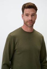 Olivengroene lange mouw shirt met een ronde hals, gladde stof en subtiele schoudernaaddetails. Model toont de vooraanzicht.